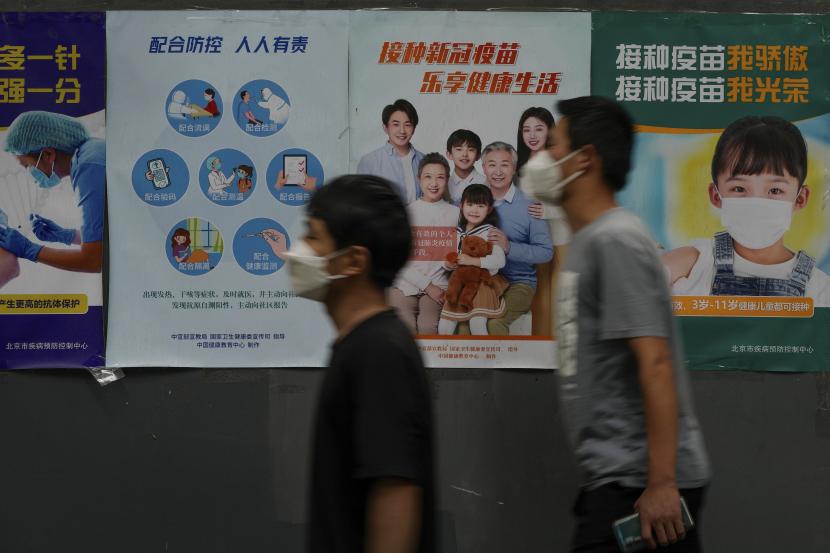 Warga yang mengenakan masker berjalan di dekat dinding yang memajang berbagai poster propaganda yang mendorong orang untuk mendapatkan vaksinasi terhadap COVID-19 di sepanjang jalan di Beijing, Kamis, 9 Juni 2022. 