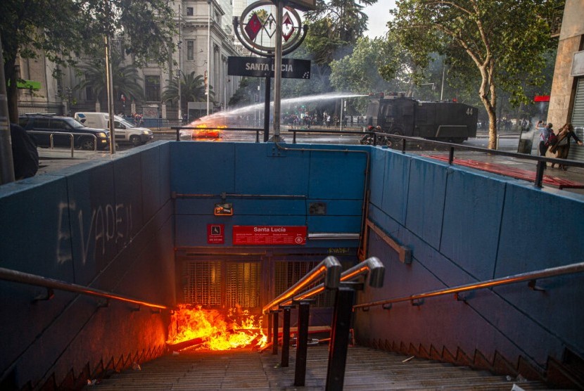 Water cannon polisi memadamkan barikade yang dibakar demonstran di stasiun kereta bawah tanah Santa Lucia di Santiago, Cile, Jumat (18/10).