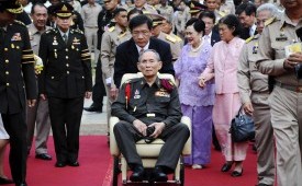 Raja Bhumibol Adulyadej didorong di kursi roda. Terlihat Ratu Sirikit mengenakan busana biru berjalan di belakangnya.