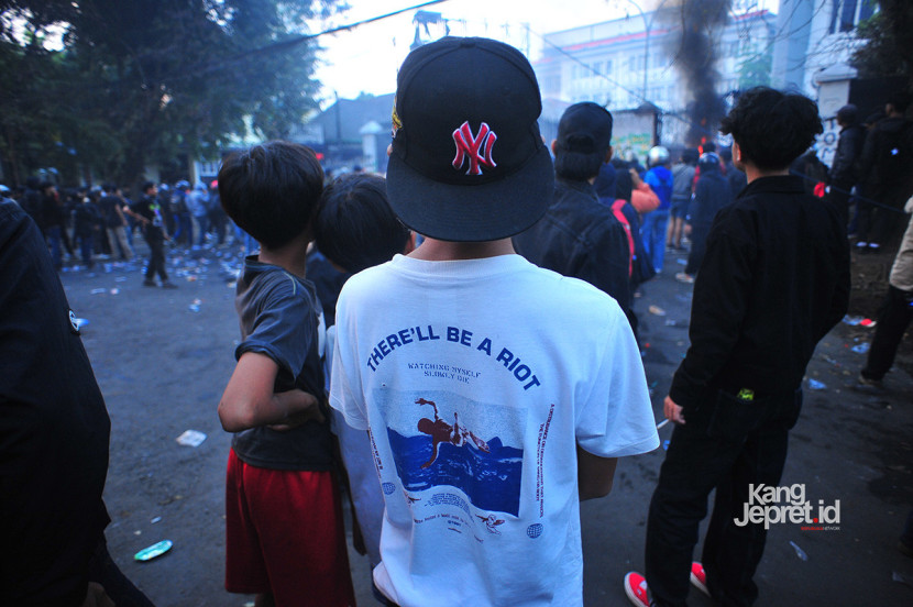 Seorang anak mengenakan kaus bertulisankan 'Akan Ada Kerusuhan' menonton aksi pembakaran oleh pengunjukrasa di gerbang Gedung DPRD Jawa Barat, Jl Diponegoro, Bandung, Senin (1/9/2025), Aksi unjukrasa beberapa hari berubah menjadi tontonan warga dan pedagang yang mencari peruntungan di keramaian massa. (KANGJEPRET.ID/YOGI ARDHI) (Nikon D3, Nikkor 20/2.8 D)