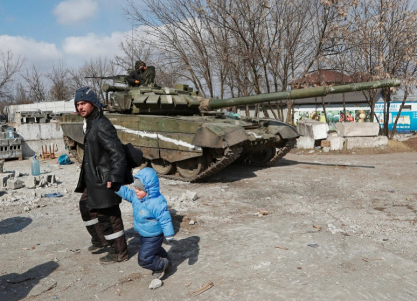 Seorang penduduk lokal berjalan dengan seorang anak melewati tank pasukan Rusia di Mauripol.