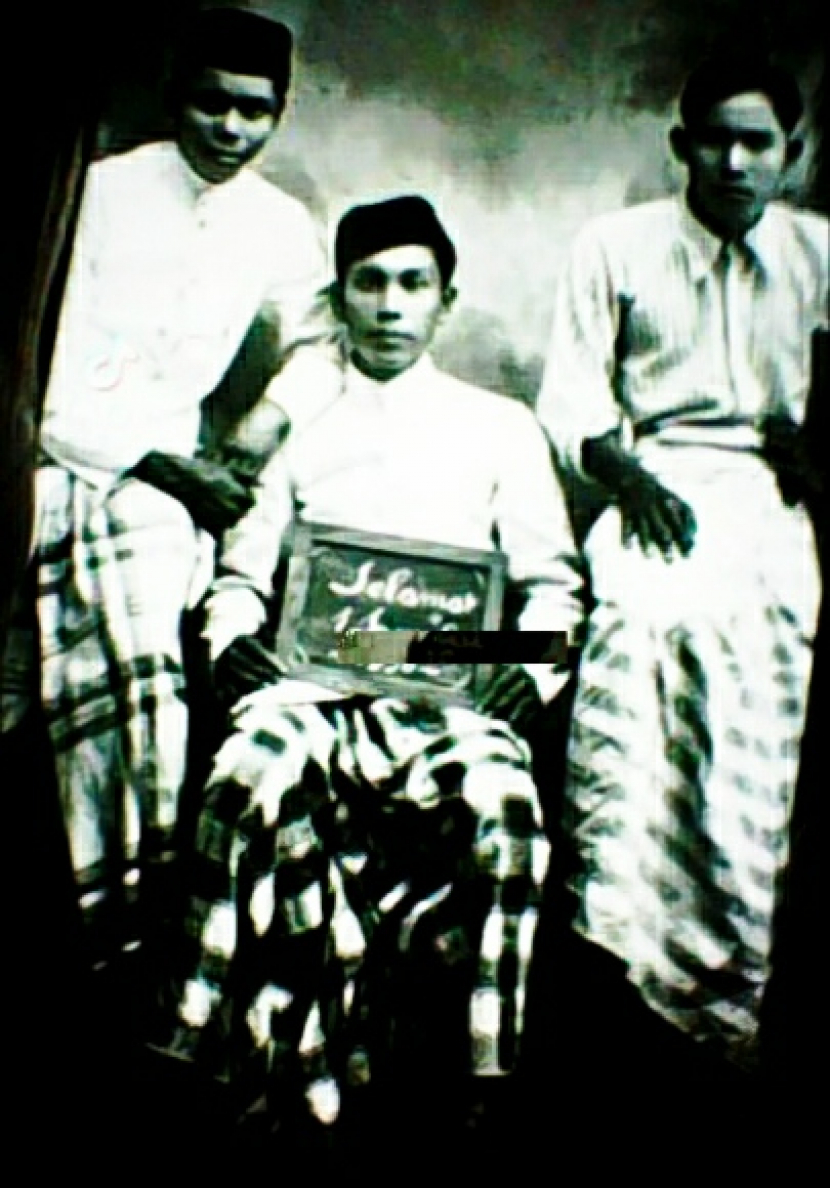 Foto tiga pria Betawi tahun 1918, ada yang memegang papan lé, papan tulis kecil, bertuliskan Selamat 1 Sjawal.
